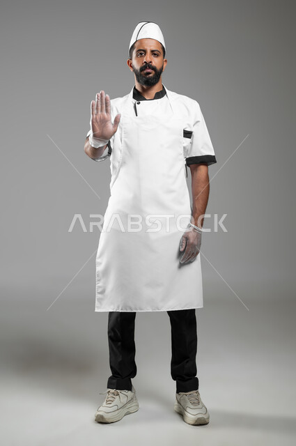 طباخ ينظر الى الكاميرا و يرفع يده للحذر من الاقتراب, بورتريه لشيف عربي سعودي خليجي يرتدي يونيفورم الطبخ يشير بيده لطلب التوقف والابتعاد، صورة الجسم بالطول الكامل, خلفيه رماديه، محتوى حساس، استخدام حساس
