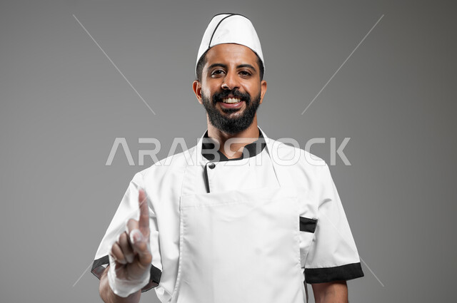 تعابير الرقم واحد, طباخ عربي خليجي سعودي يرتدي يونيفورم الطبخ ينظر الى الكاميرا بتعابير السرور، بورتريه مقرب لشيف يرفع اصبع السبابة للاعلى ويشير الى شيء ما، خلفيه رماديه، محتوى حساس، استخدام حساس