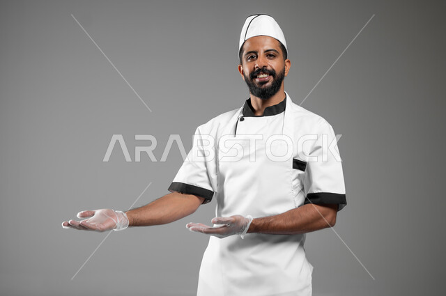 طباخ ينظر الى الكاميرا بإيماءات حمل عنصر ما ,بورتريه لشيف عربي خليجي سعودي يرتدي يونيفورم الطبخ يفتح يديه ويشير الى شيء ما , ايماءات السعاده والسرور, الفخر بالطبخ السعودي, خلفية رمادية, محتوى حساس, استخدم حساس
