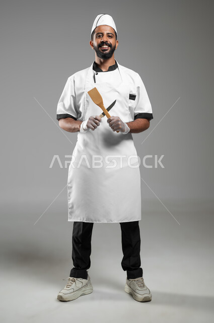 Preparing to eat food, looking at the camera with an expression of pleasure, a portrait of a smiling Saudi Arabian Gulf chef wearing a cooking uniform working in the field of cooking, a cook holding food tools in a criss-crossed manner with his hands, full-length body photo, gray background, sensitive content, sensitive use