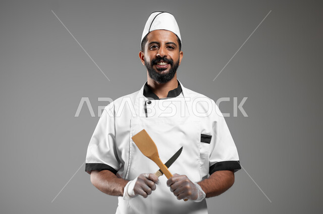 النظر الى الكاميرا بتعابير السرور, بورتريه مقرب لشيف عربي سعودي خليجي مبتسم يرتدي يونيفورم الطبخ يعمل في مجال الطهي، طباخ يحمل بيديه ادوات الطعام بشكل متقاطع، خلفيه رماديه، محتوى حساس، استخدام حساس