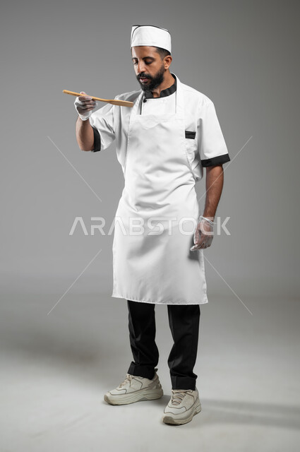 القيام في تحضير الوجبات الشهية، بورتريه لشيف سعودي عربي خليجي يرتدي يونيفورم الطبخ يعمل في مجال الطهي, طباخ يمسك بيده ملعقه خشبيه يقوم بتذوق الطعام، صوره الجسم بالطول الكامل، خلفيه رماديه، محتوى حساس, استخدام حساس