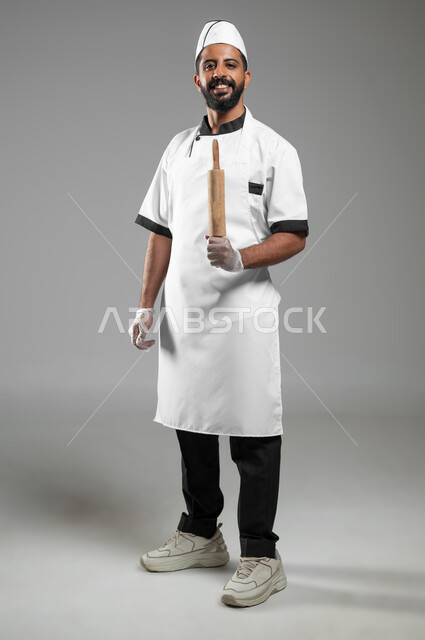 Preparing to make pastries, using manual cooking tools, a cook looking at the camera preparing to perform an operation in the kitchen, portrait of a Saudi Arabian Gulf chef wearing a cooking uniform holding a wooden mixer in his hand, full-length body photo, gray background, sensitive use, sensitive content