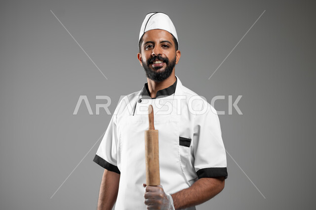 طباخ ينظر الى الكاميرا يستعد لأداء عملية في المطبخ ,استخدام أدوات الطهي اليدويه، بورتريه مقرب لشيف عربي خليجي سعودي يرتدي يونيفورم الطبخ يمسك بيده عجانة خشبية، خلفيه رماديه، استخدام حساس، محتوى حساس