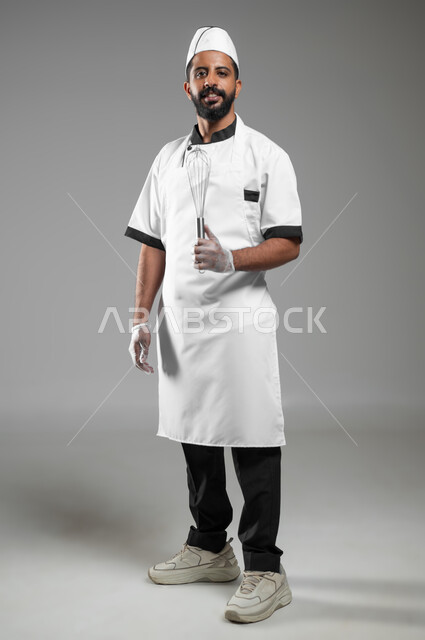 استخدام أدوات الطهي اليدويه، بورتريه لشيف عربي خليجي سعودي يرتدي يونيفورم الطبخ يمسك بيده خفاقه معدنية، طباخ ينظر الى الكاميرا يستعد لأداء عملية في المطبخ، صورة الجسم بالطول الكامل ,خلفيه رماديه، استخدام حساس، محتوى حساس