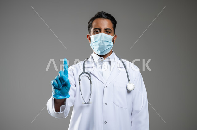ايماءات الرقم واحد ,الالتزام بالاحترازات الوقائيه ضد كوفيد 19, بورتريه مقرب لطبيب عربي سعودي خليجي يرتدي المعطف و السماعه الطبية يرفعه إصبع السبابة للاعلى، الاشارة الى مكان ما، ارتداء الكمامه لتجنب انتشار فايروس كورونا, خلفية رمادية، محتوى حساس، استخدام حساس