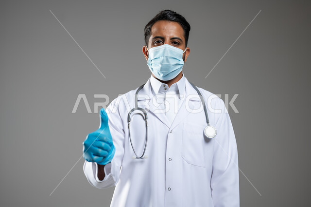 الالتزام بالاحترازات الوقائيه ضد كوفيد 19, بورتريه مقرب لطبيب عربي سعودي خليجي يرتدي المعطف و السماعه الطبية يشير بإصبع الإبهام نحو الاعلى، الرضا والقبول بجوده الخدمات الطبية، ارتداء الكمامه لتجنب انتشار فايروس كورونا, خلفية رمادية، محتوى حساس، استخدام حساس