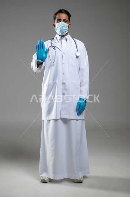 رفع اليد للحذر من الاقتراب, مفهوم إتباع الإجراءات الوقائيه لضمان السلامة من فايروس كورونا، بورتريه لطبيب عربي خليجي سعودي يرتدي المعطف الطبي ينظر الى الكاميرا و يشير بيده لطلب التوقف والابتعاد, صورة الجسم بالطول الكامل, خلفية رمادية, محتوى حساس, استخدام حساس