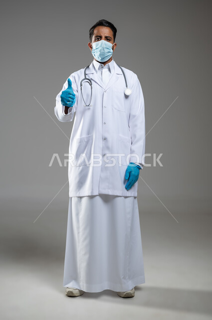 اتباع اجراءات الوقاية من كوفيد19, الرضا عن الخدمات الطبيه والرعاية الصحيه، بورتريه لطبيب عربي سعودي خليجي يرتدي المعطف الطبي  والكمامة يشير بإصبع الإبهام نحو الاعلى بايماءات الاعجاب، صورة الجسم بالطول الكامل، خلفية رمادية، محتوى حساس، استخدام حساس