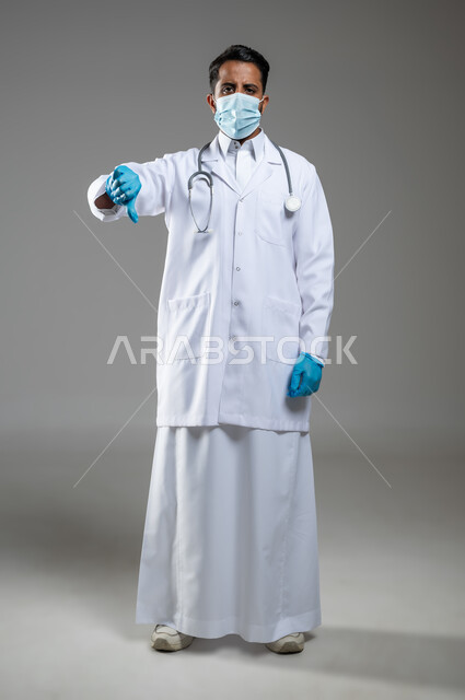 ايماءات الرفض وعدم القبول، السلامة والحماية من فايروس كورونا, بورتريه بالطول الكامل لطبيب عربي سعودي خليجي يرتدي المعطف الطبي الابيض والقفازات الطبية الزرقاء، يضع كمامة طبيه ويشير بأصبع الابهام نحو الاسفل، خلفية رمادية، محتوى حساس، استخدام حساس