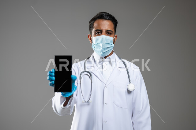 اتباع اجراءات الوقاية من كوفيد19, السلامة والحماية من فايروس كورونا, بورتريه مقرب لطبيب عربي سعودي خليجي  يرتدي المعطف الطبي والكمامة والقفازات يقوم بعرض شاشة فارغة سوداءعبر الهاتف المحمول، خلفية رمادية، محتوى حساس، استخدام حساس