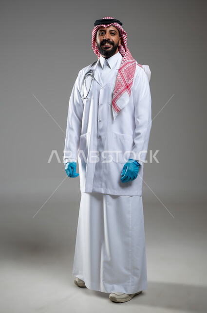 الوقوف باستقامة بايماءات الثقة بالنفس، العمل في القطاع الطبي، تقديم الخدمات الصحيه، بورتريه لطبيب عربي خليجي سعودي مبتسم يرتدي معطف الطب ينظر الى الكاميرا بتعابير السرور، صورة الجسم بالطول الكامل، خلفية رمادية، محتوى حساس، استخدام حساس