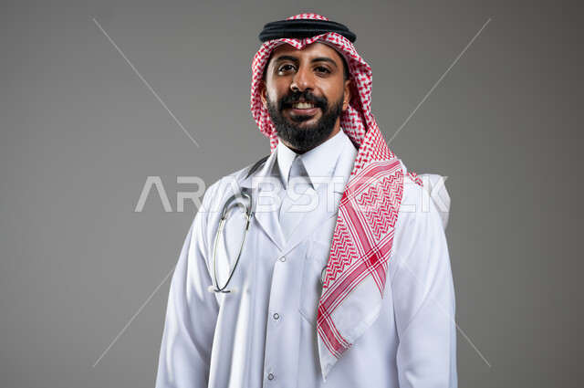 العمل في القطاع الطبي، تقديم الخدمات الصحية، بورتريه مقرب لطبيب عربي خليجي سعودي مبتسم يرتدي المعطف الطبي ينظر الى الكاميرا بتعابير السرور، خلفية رمادية، محتوى حساس، استخدام حساس