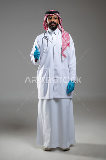 الرضا عن الخدمات الطبيه والرعاية الصحيه، إيماءات مختلفة تدل على علامات الجودة، بورتريه مقرب لطبيب عربي سعودي خليجي يرتدي المعطف الطبي والثوب التقليدي والشماغ يرفع أصبع الأبهام بتعابير الاعجاب، صورة الجسم بالطول الكامل، خلفية رمادية، محتوى حساس، استخدام حساس