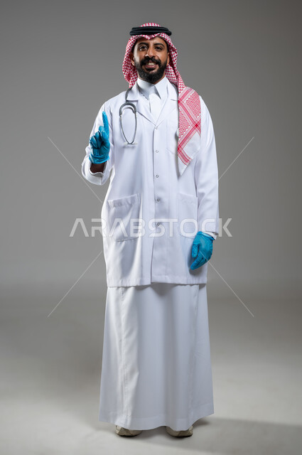 لنظر الى الكاميرا بتعابير السرور، مفهوم الطب و الرعاية الصحية، بورتريه لطبيب عربي سعودي خليجي مبتسم يرتدي المعطف الطبي والزي التقليدي والشماغ يرفع أصبع السبابة نحو الأعلى، الاشارة لشيء ما، صورة الجسم بالطول الكامل، خلفية رمادية، محتوى حساس، استخدام حساس