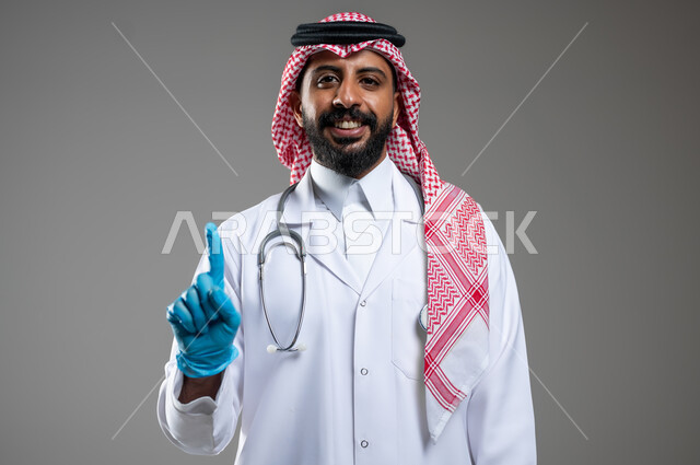 النظر الى الكاميرا بتعابير السرور، مفهوم الطب و الرعاية الصحية، بورتريه مقرب لطبيب عربي سعودي خليجي مبتسم يرتدي الثوب مع المعطف الطبي والشماغ يرفع أصبع السبابة نحو الأعلى ليشير لشيء ما، خلفية رمادية، محتوى حساس، استخدام حساس