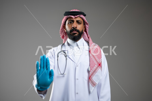 مفهوم الطب و الرعاية الصحية، النظر الى الكاميرا بتعابير الغضب والاستياء، بورتريه مقرب لطبيب عربي سعودي خليجي يرتدي الثوب مع المعطف الطبي والشماغ يشير بيده بالكامل إلى الكاميرا، خلفية رمادية،  محتوى حساس، استخدام حساس