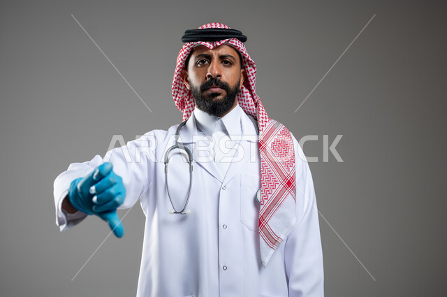 النظر الى الكاميرا بتعابير الغضب والاستياء، مفهوم الطب و الرعاية الصحية، بورتريه مقرب لطبيب عربي سعودي خليجي يرتدي الثوب مع المعطف الطبي والشماغ يشير بأصبعه نحو الاسفل بأيماءات الرفض وعدم الرضى وعدم الاعجاب، خلفية رمادية، محتوى حساس، استخدام حساس