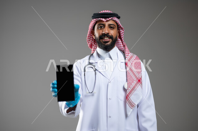 النظر الى الكاميرا بتعابير السرور، مفهوم الطب و الرعاية الصحية، بورتريه مقرب لطبيب عربي سعودي خليجي مبتسم يرتدي الثوب مع المعطف الطبي والشماغ يقوم بعرض شاشة فارغة سوداءعبر الهاتف المحمول،  خلفية رمادية، محتوى حساس، استخدام حساس