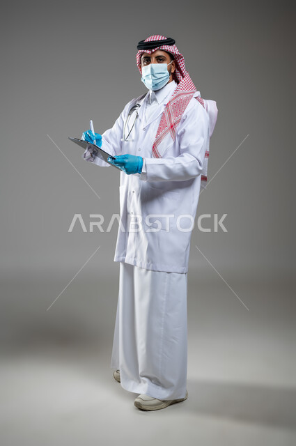 اتباع اجراءات السلامة وتقدم خدماته للمرضى, العودة بحزر واتخاذ الاحتياطات, بورتريه لطبيب عربي خليجي سعودي يضع كمامة ويرتدي المعطف طبي وقفازات يقوم بكتابة الوصفات الطبية العلاجية لكوفيد 19, الجسم بالطول الكامل, خلفية رمادية, محتوى حساس, استخدتم حساس