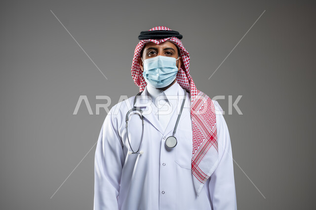 اتخاذ اجراءات للحماية من كوفيد 19, بورتريه مقرب لطبيب عربي خليجي سعودي يرتدي السماعات الطبية والكمامة للوقاية من فارس  ينظر إلى الكاميرا, يرتدي الثوب التقليدي الكمامه وسماعة طبية, خلفية رمادية, محتوى حساس, اسخدام حساس