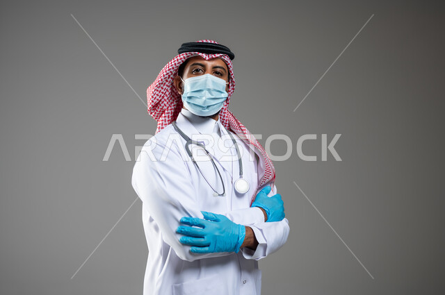 اتباع اجراءات الوقاية من كوفيد19, السلامة والحماية من فايروس كورونا,  بورتريه مقرب لطبيب عربي خليجي سعودي يرتدي الثوب التقليدي والقفازات والكمامة لضبط انتشار العدوى وسماعة طبية , خلفية رمادية, محتوى حساس, استخدام حساس