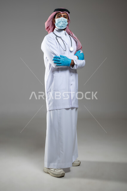 السلامة والحماية من فايروس كورونا, اتباع اجراءات الوقاية من كوفيد19, بورتريه لطبيب عربي خليجي سعودي يرتدي الثوب التقليدي والقفازات والكمامة لضبط انتشار العدوى وسماعة طبية, الجسم بالطول الكامل, خلفية رمادية, محتوى حساس, استخدام حساس