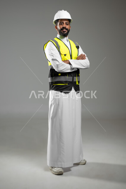 Working in the engineering sector, the concept of engineering and architectural construction, a portrait of a smiling Saudi Arabian Gulf engineer wearing a helmet and protective vest looking at the camera, standing with crossed hands, Saudi professions and jobs, full-length body, gray background, sensitive content, sensitive use