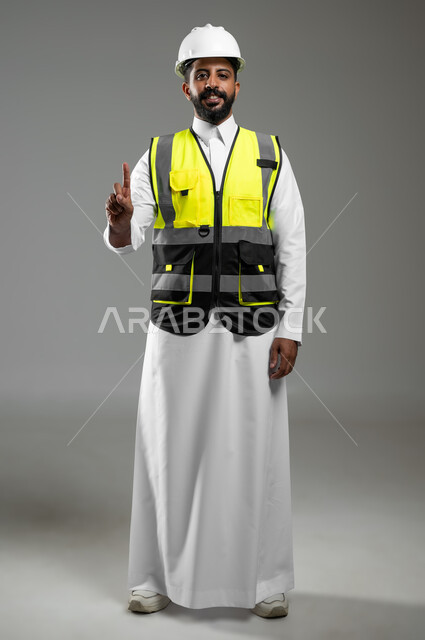 ايماءات يد تشير لرقم واحد الاشارة إلى الاعلى حركات تدل على هذا المكان,  الفخر والإعتزاز بالنفس,  بورتريه لمهندس عربي خليجي سعودي يرتدي الخوذة وسترة الحماية, لجسم بالطول الكامل, خلفية رمادية, محتوى حساس، استخدام حساس