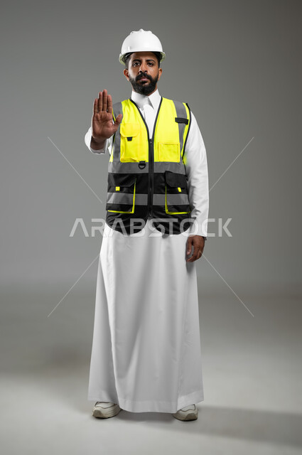 رفع اليد للحذر من الاقتراب, النظر إلى الكاميرا بايماءات الانزعاج والجدية, بورتريه لمهندس عربي خليجي سعودي يرتدي الخوذة وسترة الحماية يشير بيده لطلب التوقف والابتعاد, الجسم بالطول الكامل, خلفية رمادية, محتوى حساس, استخدام حساس