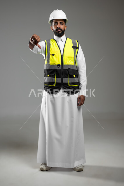 حركات تدل على القلق وعدم الموافقة والإعجاب, بورتريه لمهندس عربي سعودي خليجي يشير بإصبعه الإبهام لأسفل بإيماءات وجه ويدين مختلفة تدل على الرفض وعدم الموافقة, الجسم بالطول الكامل,  خلفية رمادي, محتوى حساس, استخدام حساس