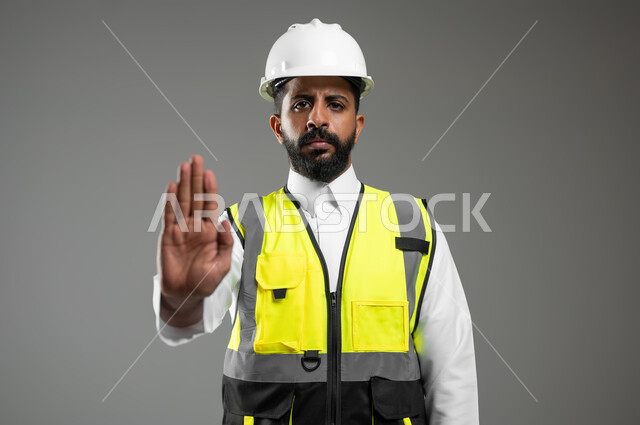 النظر إلى الكاميرا بايماءات الانزعاج والجدية, رفع اليد للحذر من الاقتراب, بورتريه مقرب لمهندس عربي خليجي سعودي يرتدي الخوذة وسترة الحماية يشير بيده لطلب التوقف والابتعاد, خلفية رمادية, محتوى حساس, استخدام حساس
