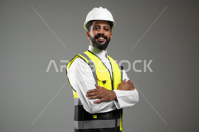 مفهوم الهندسة والانشاءات المعمارية،العمل في القطاع الهندسي،   بورتريه مقرب لمهندس عربي خليجي سعودي مبتسم يرتدي الخوذه وسترة الحماية ينظر إلى الكاميرا،   الوقوف بأيدي متقاطعه، العمل في القطاع الهندسي،  مهن ووظائف سعودية،  خلفية رمادية، محتوى حساس، استخدام حساس