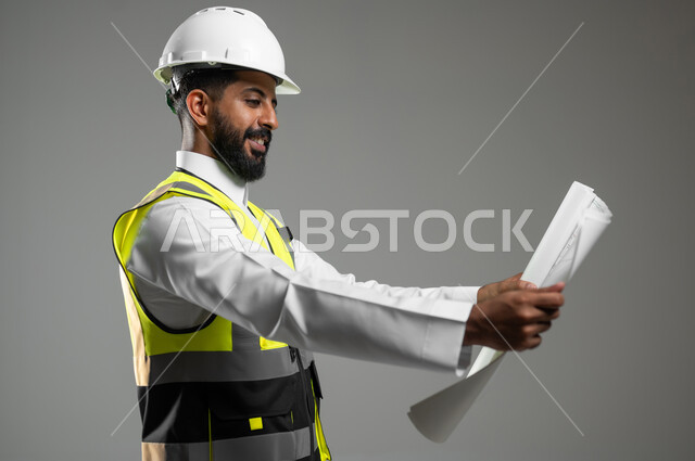 الإعمار بسواعد أبناء الوطن، مفهوم الهندسة والانشاءات المعمارية، بورتريه مقرب لمهندس عربي خليجي سعودي يرتدي خوذة وسترة الوقاية يمسك مخطط البناء وينظرإليه بفخر, المتابعة والإشراف على المشاريع الهندسية,  خلفية رمادية,  محتوى حساس،  استخدام حساس