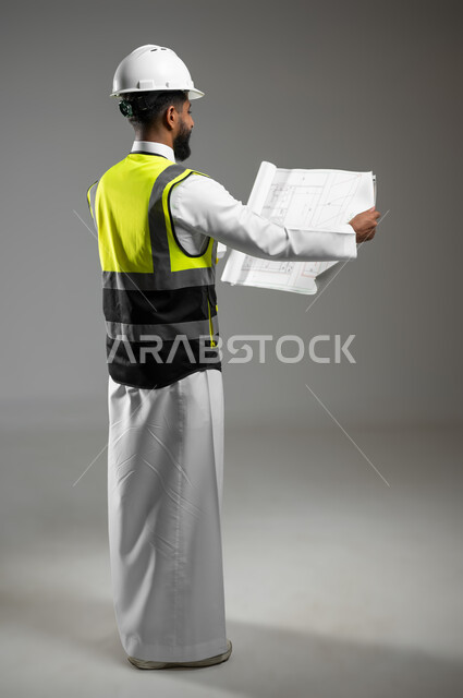 الاشراف والرقابة على المشاريع الهندسية، الاعمار بسواعد ابناء الوطن، بورتريه من الخلف لمهندس عربي خليجي سعودي يرتدي الثوب التقليدي مع خوذة وسترة الوقاية يمسك مخطط البناء وينظر إليه بفخر، الجسم بالطول الكامل، خلفية رمادية، محتوى حساس، استخدام حساس