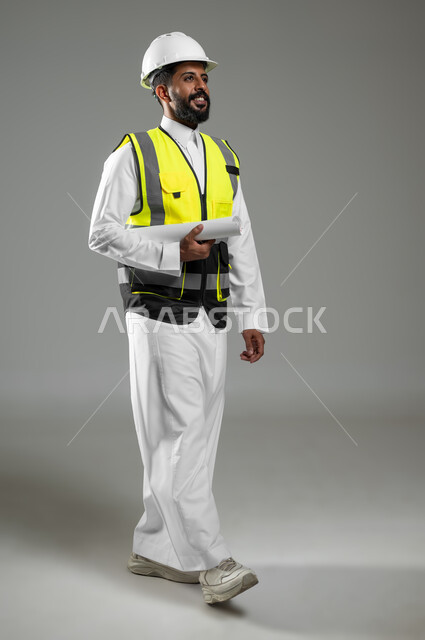 متابعة الأعمال والمشاريع، أوراق خاصة بالعمل، بورتريه لرجل خليجي عربي سعودي يرتدي الثوب التقليدي مع سترة وخوذة الحماية الخاصة بالعمل يحمل بيده مخطط البناء يمشي باتجاه الأمام، الجسم بالطول الكامل، خلفية رمادية , استخدام حساس , محتوى حساس