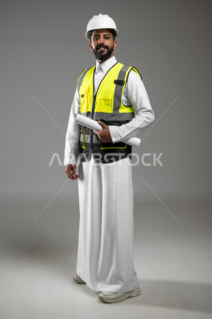 أوراق خاصة بالعمل،  بورتريه لرجل خليجي عربي سعودي يرتدي الثوب التقليدي مع سترة وخوذة الحماية الخاصة بالعمل يحمل بيده مخطط البناء، الجسم بالطول الكامل، خلفية رمادية , استخدام حساس , محتوى حساس