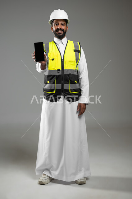عرض شاشة الهاتف المحمول الفارغة، النظر الى الكاميرا بتعابير السرور، بورتريه  لرجل خليجي عربي سعودي يرتدي الثوب التقليدي مع سترة وخوذة الحماية الخاصة بالعمل ، استخدام تقنيات حديثة، خلفية رمادية،  الجسم بالطول الكامل , محتوى حساس، استخدام حساس.