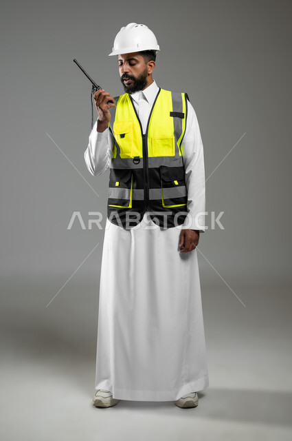 الإشراف ومتابعة العمل، بورتريه لمهندس عربي خليجي سعودي يرتدي الثوب السعودي التقليدي مع سترة وخوذة الحماية خاصة بالعمل، استخدام جهاز اتصال لاسلكي، مهن و وظائف هندسية، الجسم بالطول الكامل، خلفية رمادية , استخدام حساس , محتوى حساس