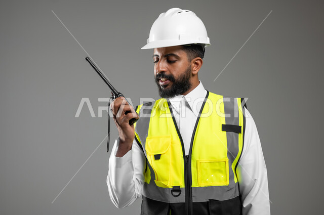 استخدام جهاز اتصال لاسلكي، بورتريه مقرب لمهندس عربي خليجي سعودي يرتدي الثوب السعودي التقليدي مع سترة وخوذة الحماية خاصة بالعمل، الإشراف ومتابعة العمل، مهن و وظائف هندسية، خلفية رمادية ,  استخدام حساس , محتوى حساس