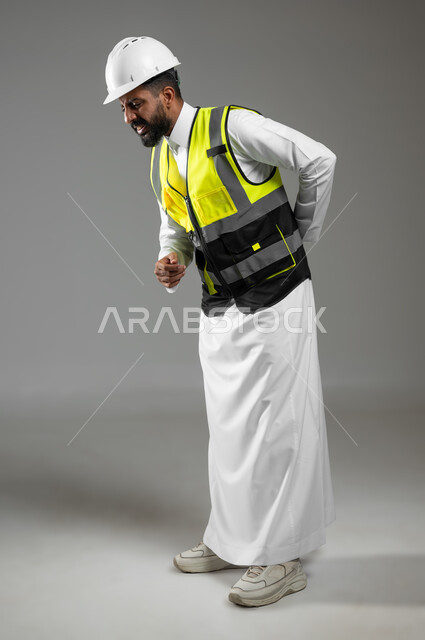ايماءات تدل على آلام الظهر والعمود الفقري , بورتريه جانبي لرجل عربي سعودي خليجي يرتدي الثوب التقليدي مع خوذة وسترة الحماية يضع يديه على ظهره يعاني من التعب والارهاق , خلفية رمادية , الجسم بالطول الكامل , استخدام حساس , محتوى حساس