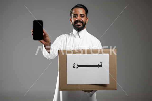 عرض شاشة الهاتف المحمول الفارغة، النظر الى الكاميرا بتعابير السرور، بورتريه مقرب لرجل خليجي عربي سعودي يرتدي الثوب التقليدي يحمل بيده صندوق التبرعات، استخدام تقنيات حديثة، خلفية رمادية، محتوى حساس، استخدام حساس.