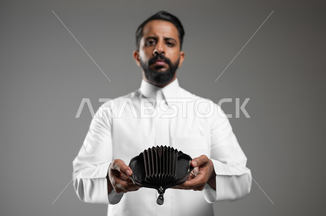 Expressions of sadness and depression, the concept of bankruptcy and not having money, a close-up portrait of the hand of a Saudi Gulf Arab young man wearing a traditional dress, opening an empty leather pocket wallet with his hand, gestures of poverty and need for money and assistance, gray background, sensitive content, sensitive use