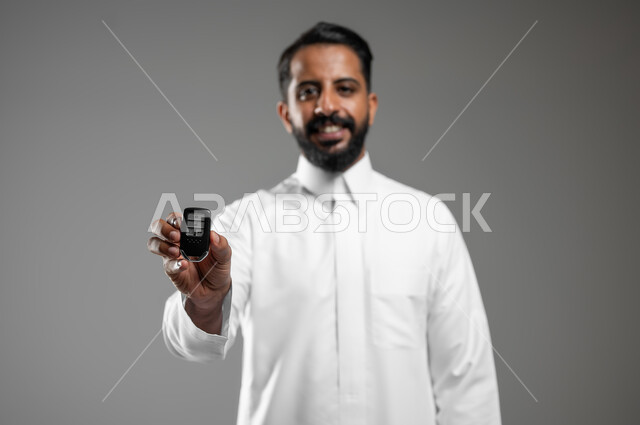ايماءات الفرح والسرور بامتلاك سيارة جديده, بورتريه لرجل عربي خليجي سعودي مبتسم يرتدي الثوب التقليدي يمسك مفتاح التحكم عن بعد, مفهوم القياده في السعوديه , خلفيه رماديه ,استخدام حساس , محتوى حساس