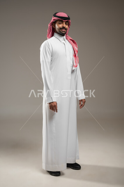 مفهوم الرجولة والقوة والثقة بالنفس، بورتريه حانبي بالطول الكامل لرجل عربي خليجي سعودي يرتدي الشماغ والثوب السعودي ينظر الى الكاميرا بإيماءات الجدية والحزم، الاهتمام بالأناقة والمظهر الخارجي، خلفية رمادية، محتوى حساس، استخدام حساس