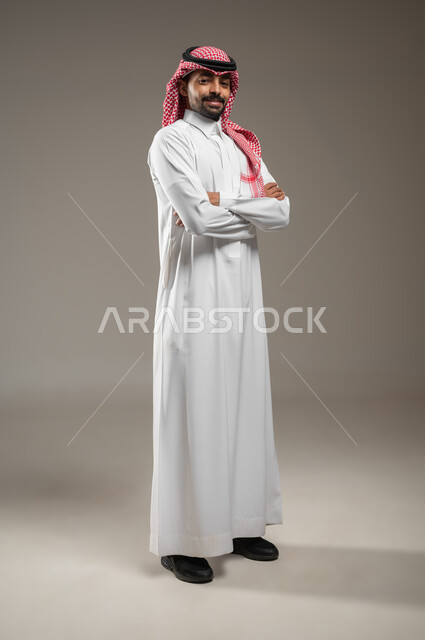 مفهوم الرجولة والقوة والثقة بالنفس، بورتريه حانبي بالطول الكامل لرجل عربي خليجي سعودي يرتدي الشماغ والثوب السعودي ينظر الى الكاميرا بإيماءات الجدية والحزم يقف مكتوف اليدين،  الاهتمام بالأناقة والمظهر الخارجي، خلفية رمادية، محتوى حساس، استخدام حساس