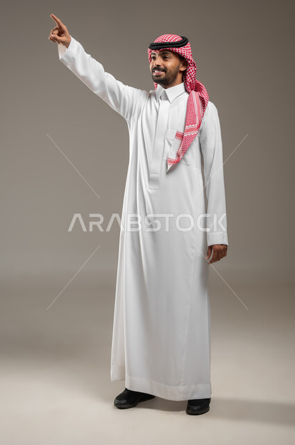 الإشارة إلى مكان ما والنظر إلى الاعلى, البشاشة والسرور وتحديد شيء معين, بورتريه بالطول الكامل لشاب عربي خليجي سعودي مبتسم يرتدي الثوب والشماغ السعودي ينظر ويشير الى الاعلى بإصبع السبابة إلى شيء ما, ايماءات العثور على مكان مجهول، خلفيه رماديه، محتوى حساس