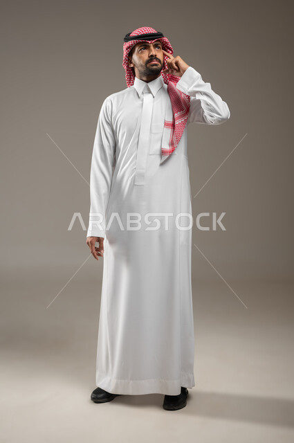 التطلع والتخطيط للمستقبل، حل المشكلات واتخاذ القرارات، بورتريه بالطول الكامل لرجل عربي سعودي خليجي يرتدي الشماغ والثوب السعودي يشير بيده نحو اذنه بإيماءات التفكير وشرود الذهن، الاستماع والتركيز، خلفية رماديه، محتوى حساس، استخدام حساس