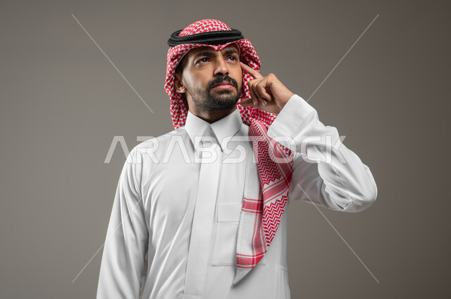 التطلع والتخطيط للمستقبل، حل المشكلات واتخاذ القرارات، بورتريه مقرب لرجل عربي سعودي خليجي يرتدي الشماغ والثوب السعودي يشير بيده نحو اذنه بإيماءات التفكير وشرود الذهن، الاستماع والتركيز، خلفية رماديه، محتوى حساس، استخدام حساس