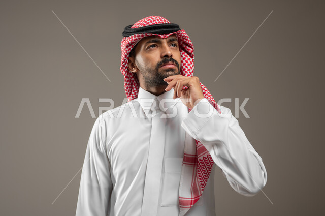 شرود الذهن والتأمل والتخطيط للمستقبل، التفكير لأيجاد حل مشكلة ما، مواجهة المشكلات والتحديات في الحياة العملية، بورتريه مقرب لرجل عربي خليجي سعودي يرتدي الزي السعودي يقف حائرا، يضع يده على ذقنه ينظر الى الأعلى بإيماءات التمعن والتركيز، خلفيه رماديه، محتوى حساس